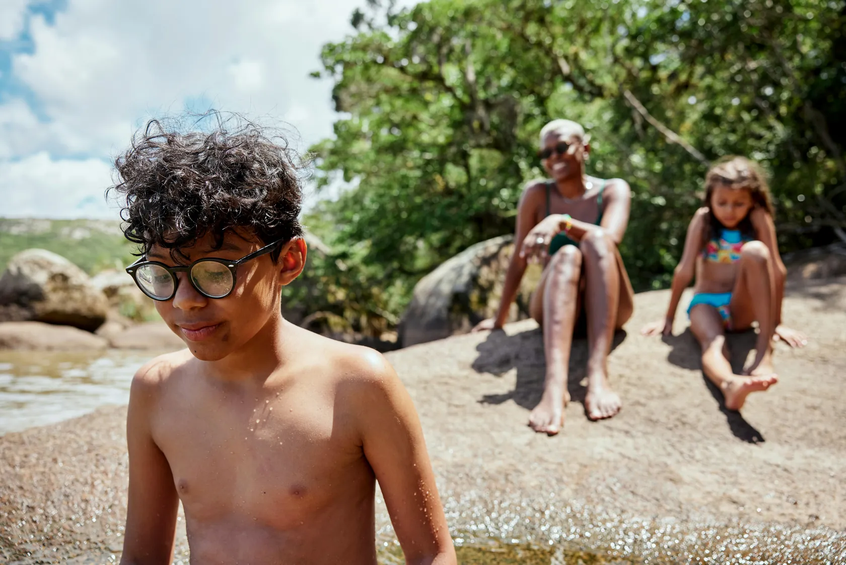 A boy in round glasses sitting on warm rocks by a river with his family